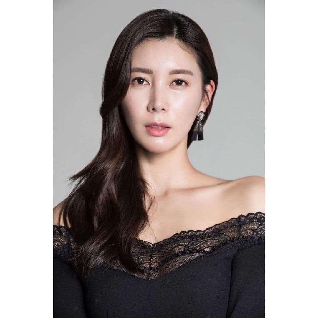Woman with long dark hair wearing a black lace off-the-shoulder top against a gray background.